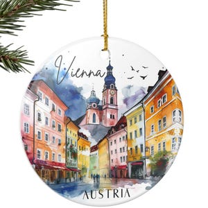 May include: Circular Christmas tree decoration with a watercolour painting of Vienna, Austria. The image depicts colourful buildings and a church with a tall spire. The word "Vienna" is in cursive, and "AUSTRIA" is printed below.
