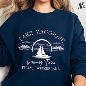 May include: Navy blue sweatshirt with a white graphic design of a sailboat on Lake Maggiore, Lombardy, Ticino, Italy, Switzerland.