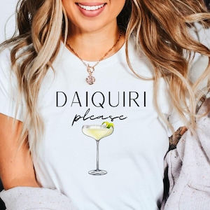 Può includere: T-shirt bianco con la scritta "DAIQUIRI please" e un'illustrazione di cocktail. Il design presenta una bevanda in stile margarita con una guarnizione di lime. La maglietta è un capo casual.