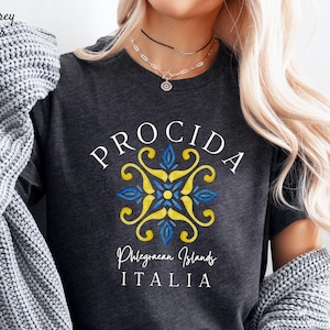 May include: Dark grey heather t-shirt with the word "PROCIDA" in white, a blue and yellow floral design, and the text "Phlegraean Islands ITALIA". The shirt is a crew neck style.