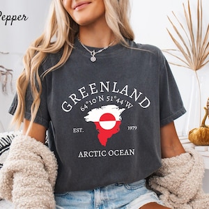 Puede incluir: Camiseta gris oscuro con un gráfico de Groenlandia. El diseño incluye la palabra "Greenland", coordenadas, un contorno del mapa con la bandera de Groenlandia y el texto "Arctic Ocean". La camiseta es de estilo casual y de manga corta.