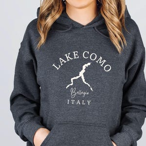 May include: Dark heather gray hoodie with a front pocket and a hood. The white text on the front reads "Lake Como" above a white outline of the lake and the word "Bellagio" and "ITALY".