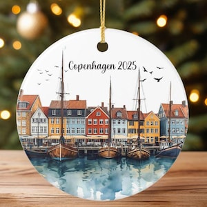 Pode incluir: Um enfeite de Natal redondo com uma pintura em aquarela de Copenhague, Dinamarca. A imagem apresenta edifícios coloridos à beira-mar com barcos e o texto "Copenhagen 2025". O enfeite é pendurado por um fio dourado.