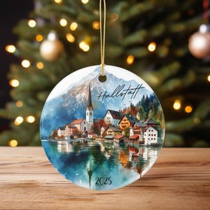 May include: A circular Christmas bauble with a watercolour depiction of Hallstatt, Austria, set against mountains and a lake reflection. It has a gold hanging string and the year 2025 printed at the bottom.
