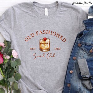 Puede incluir: Camiseta gris con el texto "Old Fashioned Est 1880 Social Club" en letras marrones. Un dibujo de un c&oacute;ctel old fashioned con una cereza como adorno est&aacute; por encima del texto.