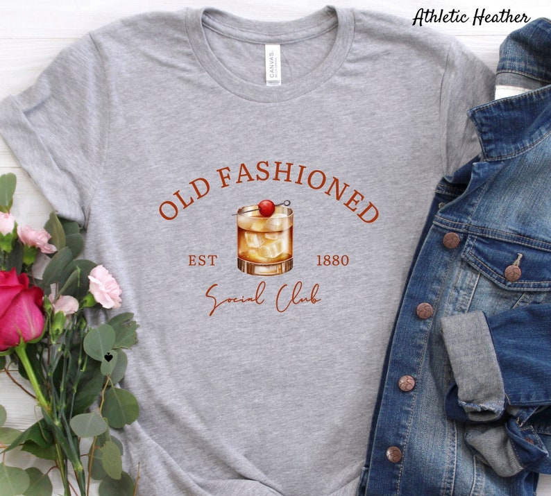 Puede incluir: Camiseta gris con el texto "Old Fashioned Est 1880 Social Club" en letras marrones. Un dibujo de un c&oacute;ctel old fashioned con una cereza como adorno est&aacute; por encima del texto.