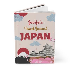 Op de afbeelding: Een hardcover reisdagboek getiteld "Jennifer's Travel Journal JAPAN". De omslag toont een rode zon, de Fuji, een Japanse burcht, roze kersenbloesems en gestileerde wolken. Het dagboek is crèmekleurig.