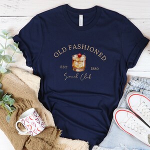 Puede incluir: Una camiseta azul marino con un gr&aacute;fico dorado de un c&oacute;ctel old fashioned con una cereza en la parte superior. El gr&aacute;fico tambi&eacute;n incluye el texto "Old Fashioned Est 1880 Social Club".