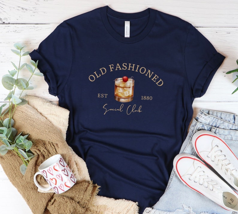 Puede incluir: Una camiseta azul marino con un gr&aacute;fico dorado de un c&oacute;ctel old fashioned con una cereza en la parte superior. El gr&aacute;fico tambi&eacute;n incluye el texto "Old Fashioned Est 1880 Social Club".