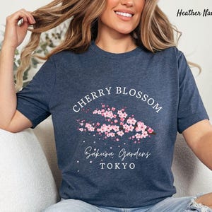 Puede incluir: Camiseta azul marino jaspeado con un dise&ntilde;o de flor de cerezo. La camiseta muestra las palabras "Cherry Blossom" sobre una rama de flores rosas. Debajo de las flores, la camiseta dice "Sakura Gardens TOKYO". La camiseta es de un material suave.