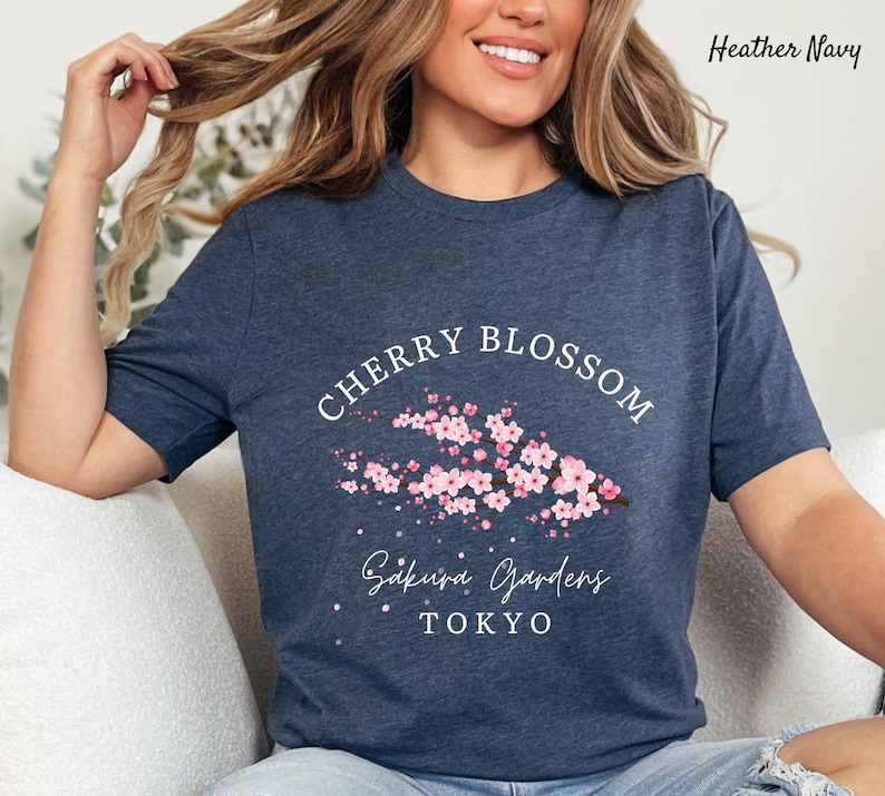 Puede incluir: Camiseta azul marino jaspeado con un dise&ntilde;o de flor de cerezo. La camiseta muestra las palabras "Cherry Blossom" sobre una rama de flores rosas. Debajo de las flores, la camiseta dice "Sakura Gardens TOKYO". La camiseta es de un material suave.