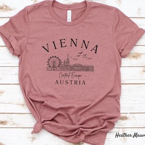 May include: Heather mauve t-shirt featuring a black graphic design of the Vienna skyline, including the Ferris wheel, St. Stephen's Cathedral, and the Hofburg Palace. The text "VIENNA", "Central Europe", and "AUSTRIA" is also included in the design.
