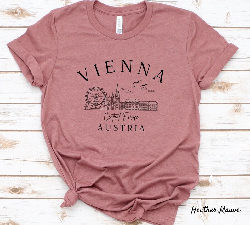May include: Heather mauve t-shirt featuring a black graphic design of the Vienna skyline, including the Ferris wheel, St. Stephen's Cathedral, and the Hofburg Palace. The text "VIENNA", "Central Europe", and "AUSTRIA" is also included in the design.