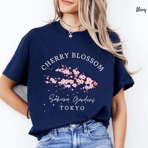Puede incluir: Camiseta azul marino con un dise&ntilde;o de "Cherry Blossom". El gr&aacute;fico presenta flores de cerezo rosas y el texto "Sakura Gardens Tokyo" en blanco. Una camiseta casual de manga corta.