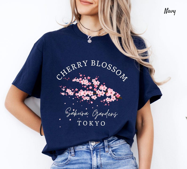 Puede incluir: Camiseta azul marino con un dise&ntilde;o de "Cherry Blossom". El gr&aacute;fico presenta flores de cerezo rosas y el texto "Sakura Gardens Tokyo" en blanco. Una camiseta casual de manga corta.