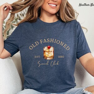 Puede incluir: Una camiseta azul marino jaspeada con un gr&aacute;fico de un c&oacute;ctel old fashioned con una cereza en la parte superior. El texto "Old Fashioned" est&aacute; por encima del c&oacute;ctel y "Est 1880 Social Club" est&aacute; por debajo del c&oacute;ctel.