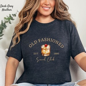 Puede incluir: Camiseta de color gris oscuro jaspeado con un gr&aacute;fico dorado de un c&oacute;ctel old fashioned con una cereza en la parte superior. El texto "Old Fashioned" est&aacute; en un c&iacute;rculo dorado sobre el c&oacute;ctel. El texto "Est 1880" est&aacute; debajo del c&oacute;ctel y el texto "Social Club" est&aacute; debajo de ese.