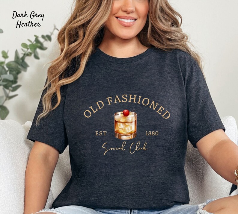 Puede incluir: Camiseta de color gris oscuro jaspeado con un gr&aacute;fico dorado de un c&oacute;ctel old fashioned con una cereza en la parte superior. El texto "Old Fashioned" est&aacute; en un c&iacute;rculo dorado sobre el c&oacute;ctel. El texto "Est 1880" est&aacute; debajo del c&oacute;ctel y el texto "Social Club" est&aacute; debajo de ese.