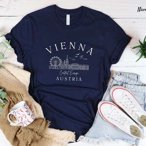 May include: Navy blue t-shirt featuring a white graphic of the Vienna skyline, including the Ferris wheel, St. Stephen's Cathedral, and the Hofburg Palace. The text "VIENNA", "Central Europe", and "AUSTRIA" is also included in the graphic.