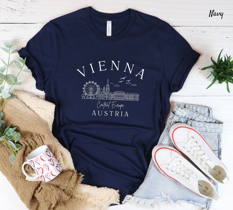 May include: Navy blue t-shirt featuring a white graphic of the Vienna skyline, including the Ferris wheel, St. Stephen's Cathedral, and the Hofburg Palace. The text "VIENNA", "Central Europe", and "AUSTRIA" is also included in the graphic.