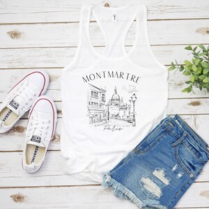 May include: White tank top featuring a black and white illustration of a Parisian street scene with the text "Montmartre Paris".