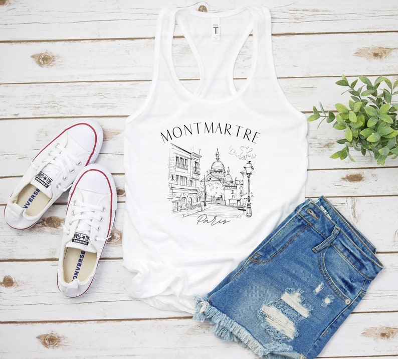 May include: White tank top featuring a black and white illustration of a Parisian street scene with the text "Montmartre Paris".