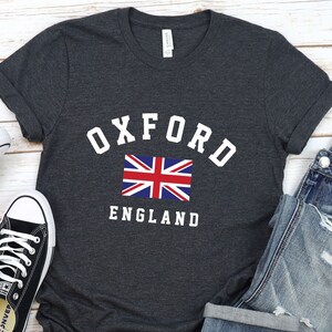 Puede incluir: Una camiseta gris oscuro con el texto "OXFORD ENGLAND" en letras blancas. Una peque&ntilde;a bandera del Reino Unido est&aacute; centrada sobre el texto.