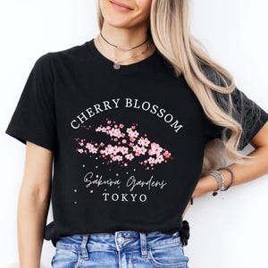 Puede incluir: Camiseta negra con un dise&ntilde;o de flor de cerezo. El dise&ntilde;o incluye el texto "Cherry Blossom", "Sakura Gardens" y "Tokyo", junto con una ilustraci&oacute;n floral rosa. La camiseta est&aacute; hecha de un material suave.