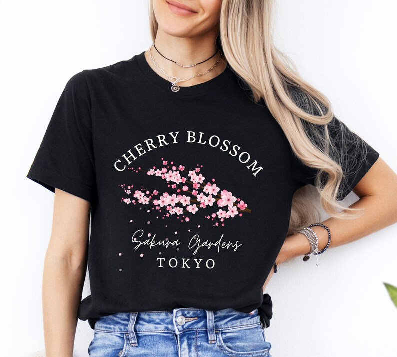 Puede incluir: Camiseta negra con un dise&ntilde;o de flor de cerezo. El dise&ntilde;o incluye el texto "Cherry Blossom", "Sakura Gardens" y "Tokyo", junto con una ilustraci&oacute;n floral rosa. La camiseta est&aacute; hecha de un material suave.