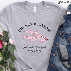 Puede incluir: Una camiseta gris claro con las palabras "CHERRY BLOSSOM" sobre una rama de flores de cerezo rosas. Debajo de las flores, el texto dice "Sakura Gardens TOKYO". La camiseta es de un material suave.