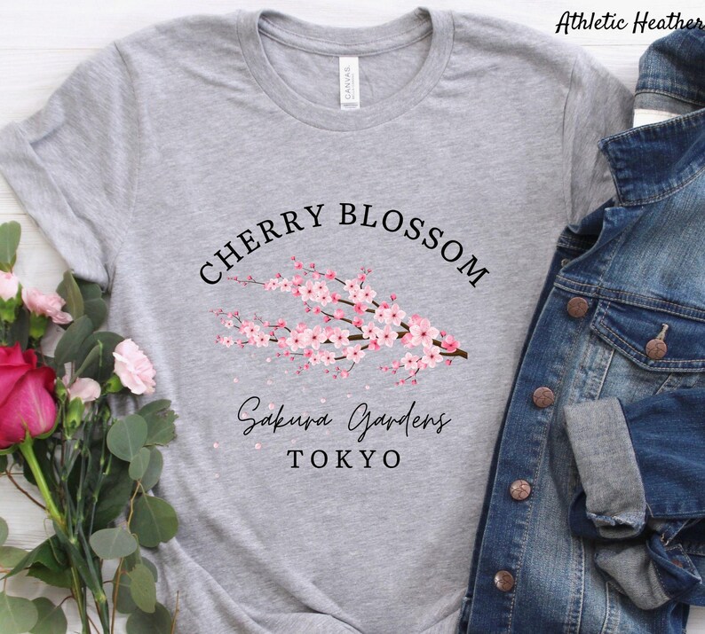 Puede incluir: Una camiseta gris claro con las palabras "CHERRY BLOSSOM" sobre una rama de flores de cerezo rosas. Debajo de las flores, el texto dice "Sakura Gardens TOKYO". La camiseta es de un material suave.