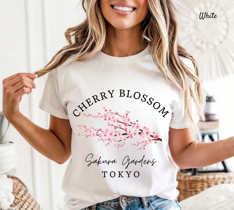 Puede incluir: Camiseta blanca con un dise&ntilde;o de flor de cerezo. El dise&ntilde;o presenta una rama con flores de cerezo rosas, las palabras "Cherry Blossom" en una fuente curva y "Sakura Gardens TOKYO" en una fuente cursiva.