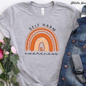 Pode incluir: Uma camiseta cinza com um design de arco-íris laranja e o texto "Self Harm Awareness".