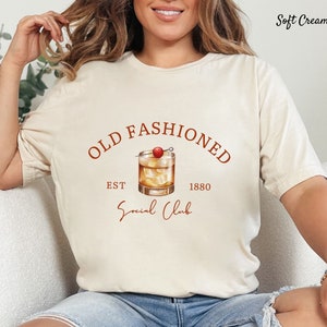 Puede incluir: Una camiseta blanca con un gr&aacute;fico marr&oacute;n de un vaso de c&oacute;ctel con una cereza en la parte superior. El texto "Old Fashioned" est&aacute; por encima del vaso, y "Est. 1880" est&aacute; por debajo del vaso. El texto "Social Club" est&aacute; escrito en cursiva debajo del vaso.