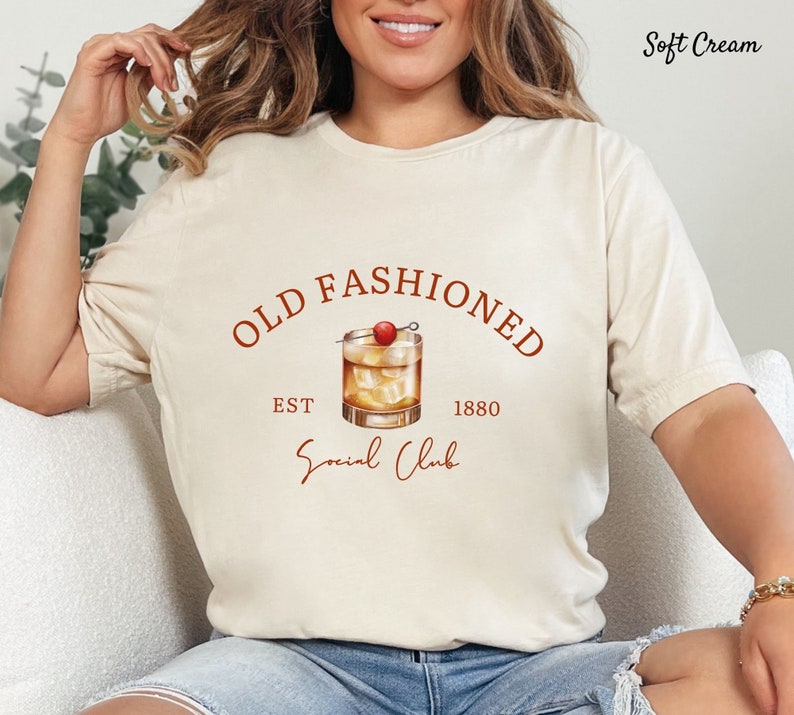 Puede incluir: Una camiseta blanca con un gr&aacute;fico marr&oacute;n de un vaso de c&oacute;ctel con una cereza en la parte superior. El texto "Old Fashioned" est&aacute; por encima del vaso, y "Est. 1880" est&aacute; por debajo del vaso. El texto "Social Club" est&aacute; escrito en cursiva debajo del vaso.