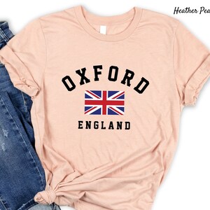 Puede incluir: Una camiseta de color melocot&oacute;n con un gr&aacute;fico negro de la palabra "OXFORD" en un arco sobre una bandera del Reino Unido y la palabra "ENGLAND" debajo de la bandera.