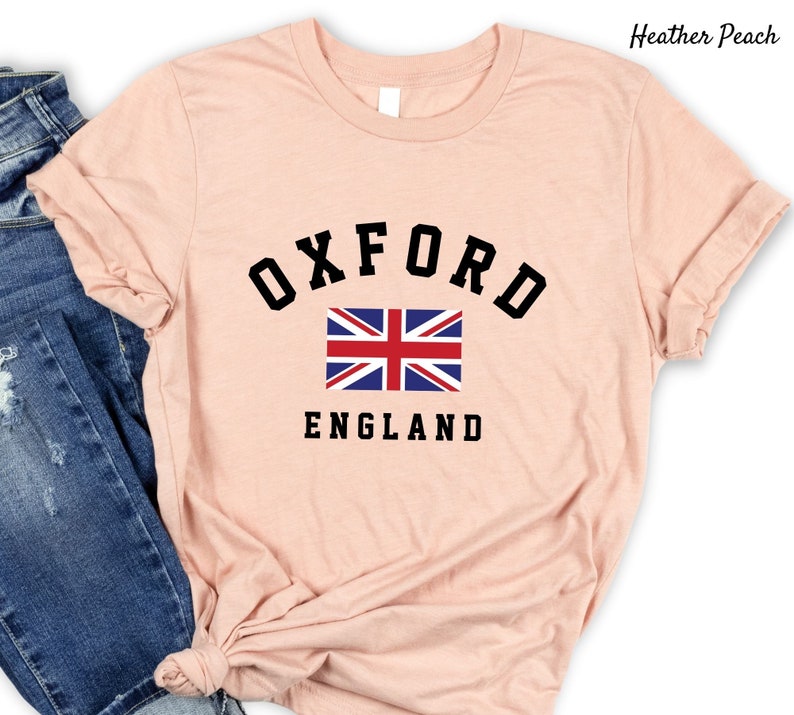 Puede incluir: Una camiseta de color melocot&oacute;n con un gr&aacute;fico negro de la palabra "OXFORD" en un arco sobre una bandera del Reino Unido y la palabra "ENGLAND" debajo de la bandera.