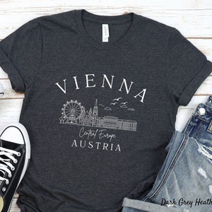 May include: Dark grey heather t-shirt with a white graphic design of the Vienna skyline, including the Ferris wheel, St Stephen's Cathedral, and the text "Vienna, Central Europe, Austria".