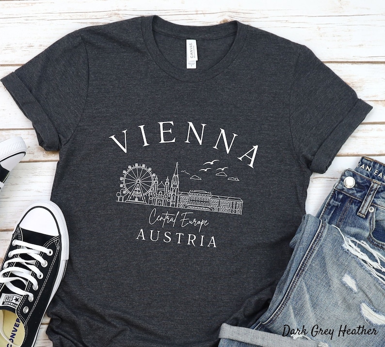 May include: Dark grey heather t-shirt with a white graphic design of the Vienna skyline, including the Ferris wheel, St Stephen's Cathedral, and the text "Vienna, Central Europe, Austria".