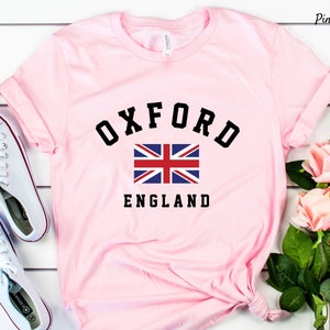 Puede incluir: Camiseta rosa con un gr&aacute;fico negro del texto "OXFORD" en una fuente universitaria, una bandera del Reino Unido y el texto "ENGLAND".