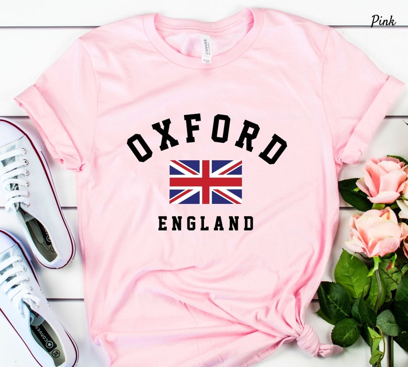 Puede incluir: Camiseta rosa con un gr&aacute;fico negro del texto "OXFORD" en una fuente universitaria, una bandera del Reino Unido y el texto "ENGLAND".