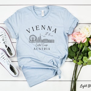 May include: Light blue t-shirt featuring a black graphic of the Vienna skyline, including the Ferris wheel, St. Stephen's Cathedral, and the Hofburg Palace. The text "VIENNA", "Central Europe", and "AUSTRIA" is printed on the shirt.