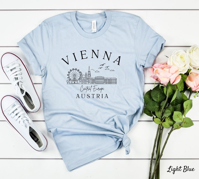 May include: Light blue t-shirt featuring a black graphic of the Vienna skyline, including the Ferris wheel, St. Stephen's Cathedral, and the Hofburg Palace. The text "VIENNA", "Central Europe", and "AUSTRIA" is printed on the shirt.