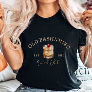 Puede incluir: Camiseta negra con texto dorado que dice "Old Fashioned Est. 1880 Social Club". Un dibujo de un c&oacute;ctel old fashioned con una cereza est&aacute; en el centro del texto.
