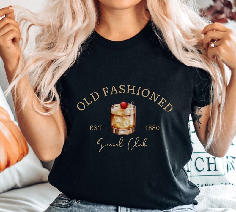 Puede incluir: Camiseta negra con texto dorado que dice "Old Fashioned Est. 1880 Social Club". Un dibujo de un c&oacute;ctel old fashioned con una cereza est&aacute; en el centro del texto.