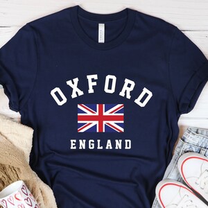 Puede incluir: Una camiseta azul marino con texto blanco que dice "Oxford England" y una peque&ntilde;a bandera del Reino Unido.