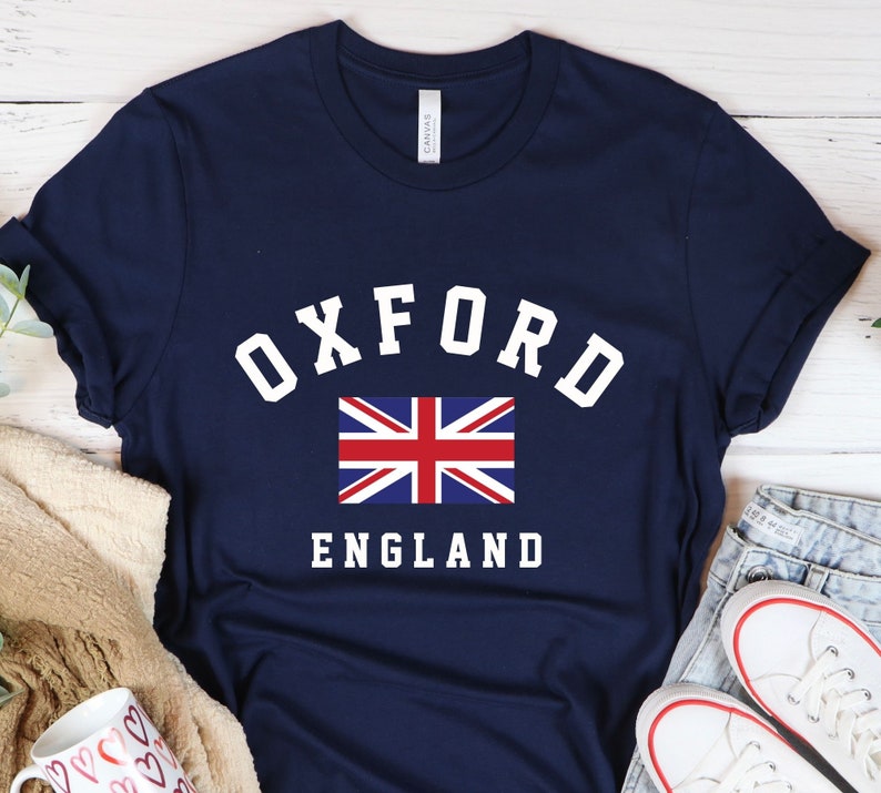 Puede incluir: Una camiseta azul marino con texto blanco que dice "Oxford England" y una peque&ntilde;a bandera del Reino Unido.