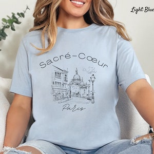 K&ouml;nnte beinhalten: Hellblaues T-Shirt mit einer Schwarz-Wei&szlig;-Skizze der Basilika Sacr&eacute;-C&oelig;ur in Paris. Die W&ouml;rter "Sacr&eacute;-Coeur" und "Paris" stehen &uuml;ber und unter der Illustration. Das T-Shirt hat eine hellblaue Farbe.