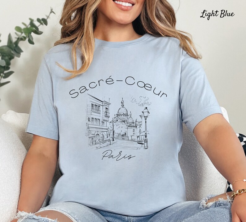 K&ouml;nnte beinhalten: Hellblaues T-Shirt mit einer Schwarz-Wei&szlig;-Skizze der Basilika Sacr&eacute;-C&oelig;ur in Paris. Die W&ouml;rter "Sacr&eacute;-Coeur" und "Paris" stehen &uuml;ber und unter der Illustration. Das T-Shirt hat eine hellblaue Farbe.