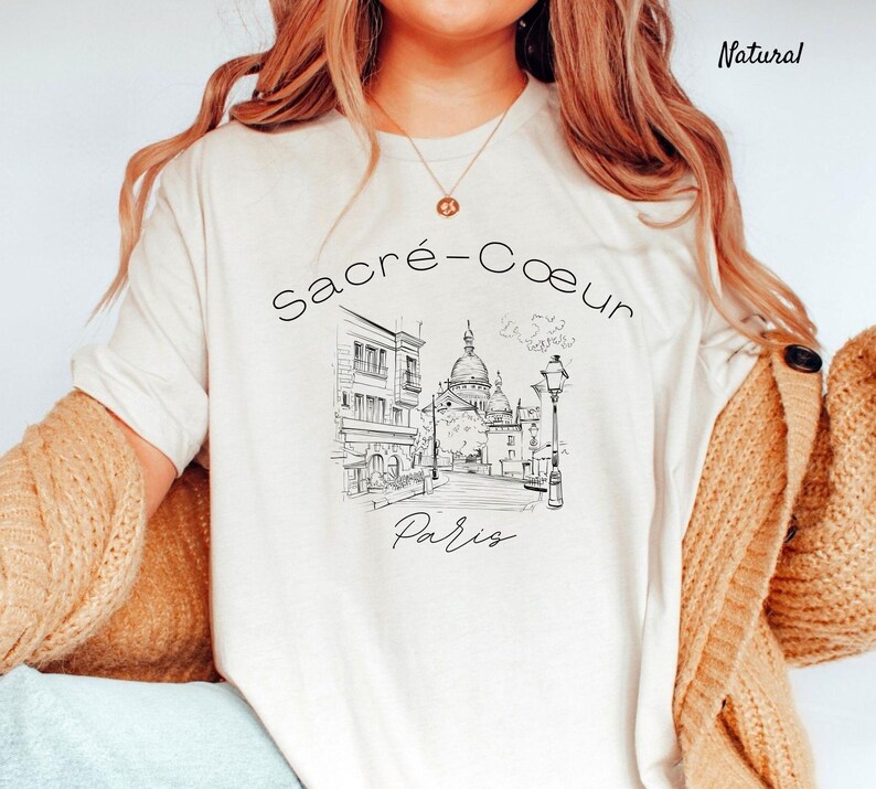 K&ouml;nnte beinhalten: Cremefarbenes T-Shirt mit einer Schwarz-Wei&szlig;-Skizze der Basilika Sacr&eacute;-C&oelig;ur in Paris. Der Text "Sacr&eacute;-Coeur Paris" ist &uuml;ber und unter der Illustration angeordnet. Das Shirt hat eine lockere Passform und kurze &Auml;rmel.